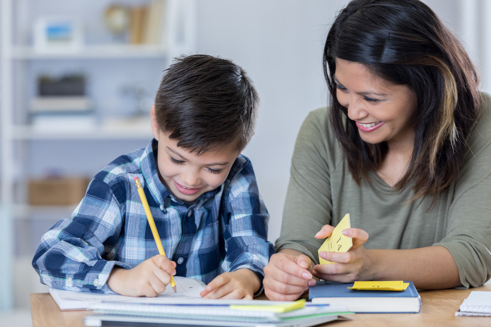 Die Mutter lächelt ihren Sohn stolz an, während er seine Mathehausaufgaben macht. Er schreibt in ein Notizbuch. Die Mutter hält Mathe-Lernkarten in der Hand.