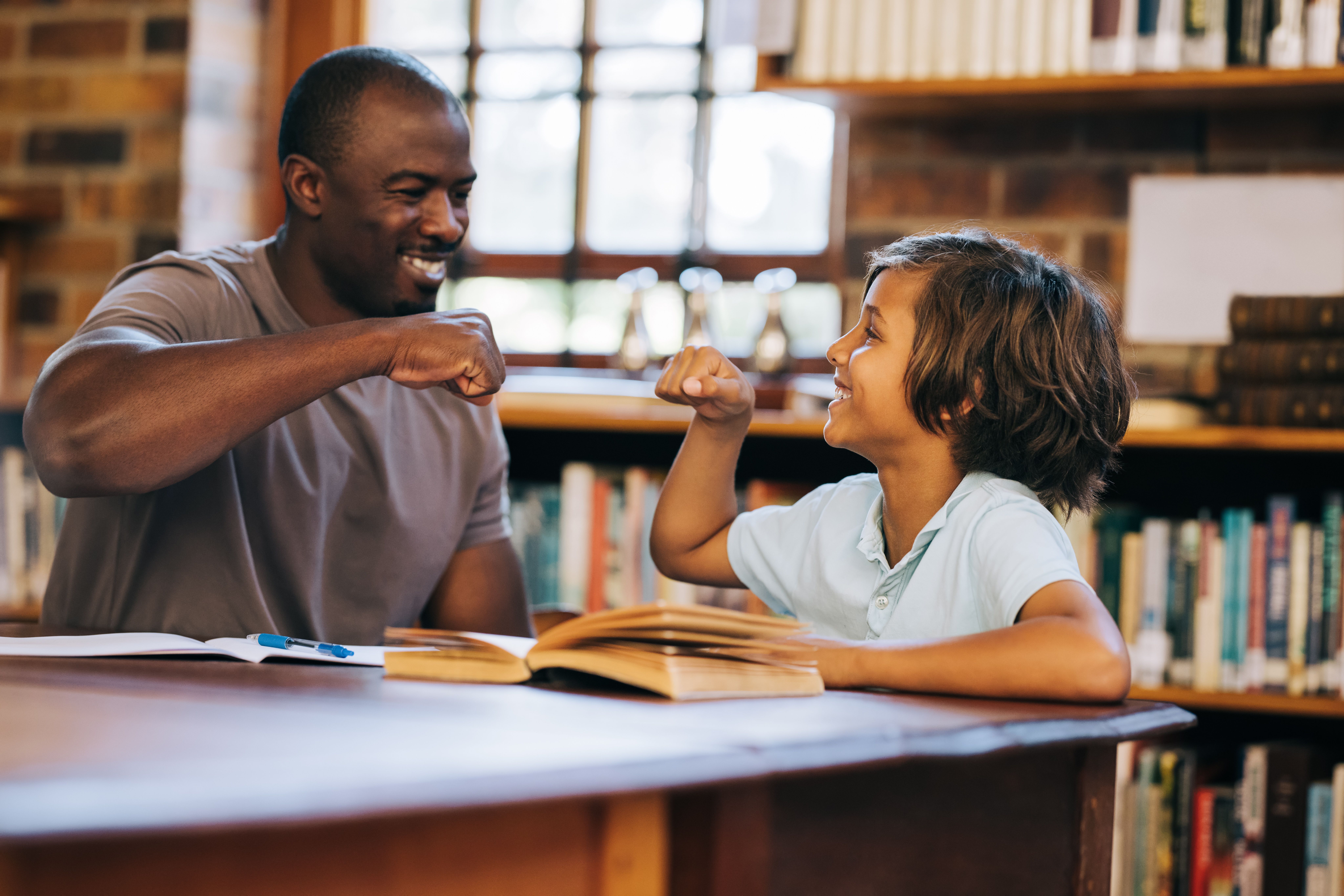 Lehrer und Schüler geben sich in einer Bibliothek ein High Five. Grundschüler im Einzelunterricht mit seiner Lehrerin. Nachhilfeunterricht und Schülerbetreuung in einer Grundschule.